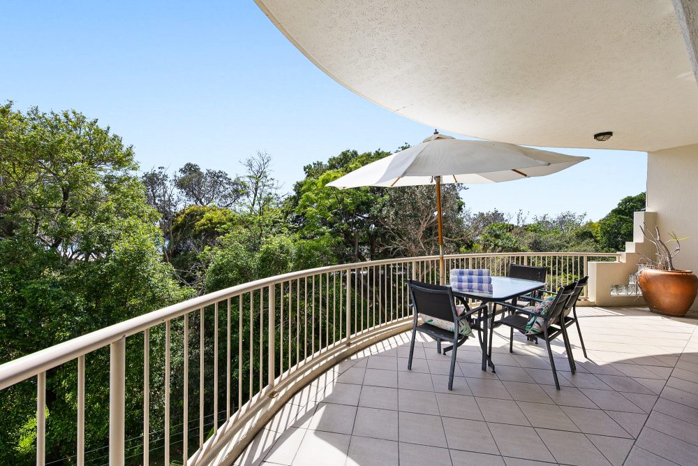 Curved balcony with outdoor dining table seating six under a large umbrella, overlooking treetops.