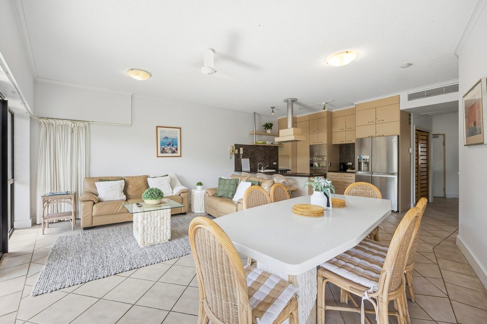 Open-plan living area with leather sofas, a dining table seating six and a kitchen with stainless steel appliances behind at Mirra Chana Beachfront Apartments Mooloolaba.