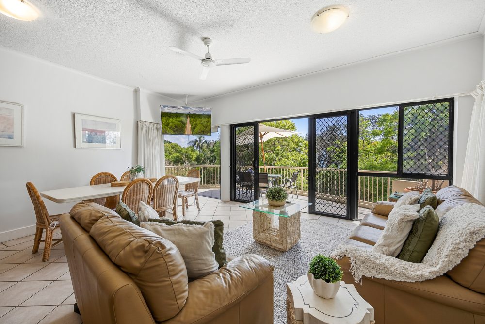 Living and dining area with leather sofa seating around a stone coffee table, opening onto a balcony surrounded by trees, plus a dining table for six.